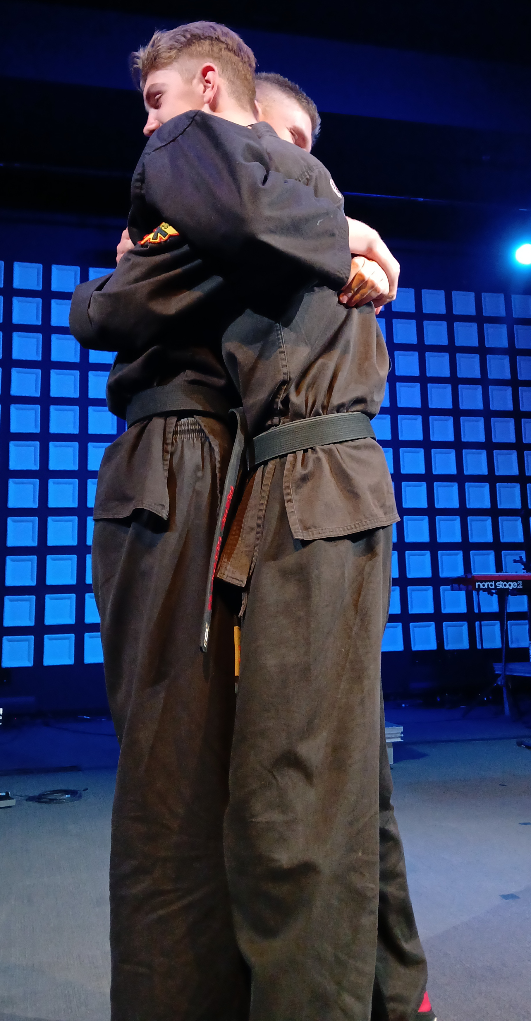 Two students embracing in black gis under blue stage lighting — the heart of Ohana